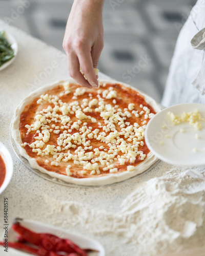 Unbaked pizza and ingredients on table. Process of making pizza. Pizzaiollo makes pizza toppings. Hand of cook in frame. Pizza with cheese and tomato sauce. Top view. Close-up. Soft focus.