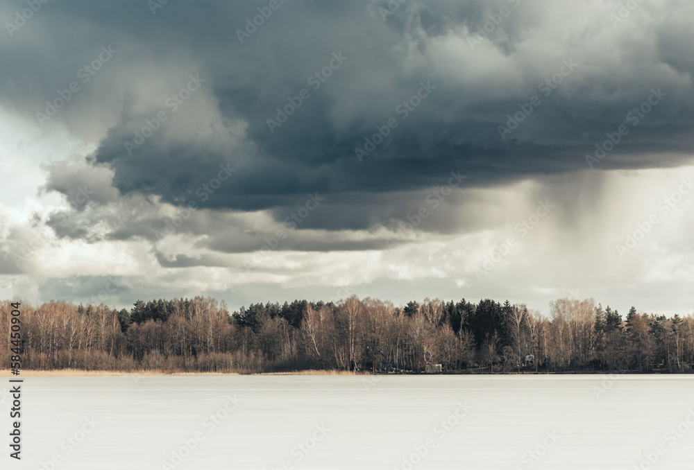 Beautiful early spring landscape with frozen lake, forest on the horizon and dramatic sky with grey clouds and snowfall in the distance, storm coming