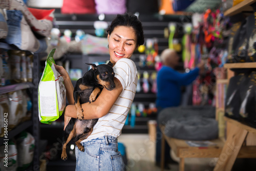 Asian woman with little dog in hands standing in salesroom of pet shop and choosing dog food.