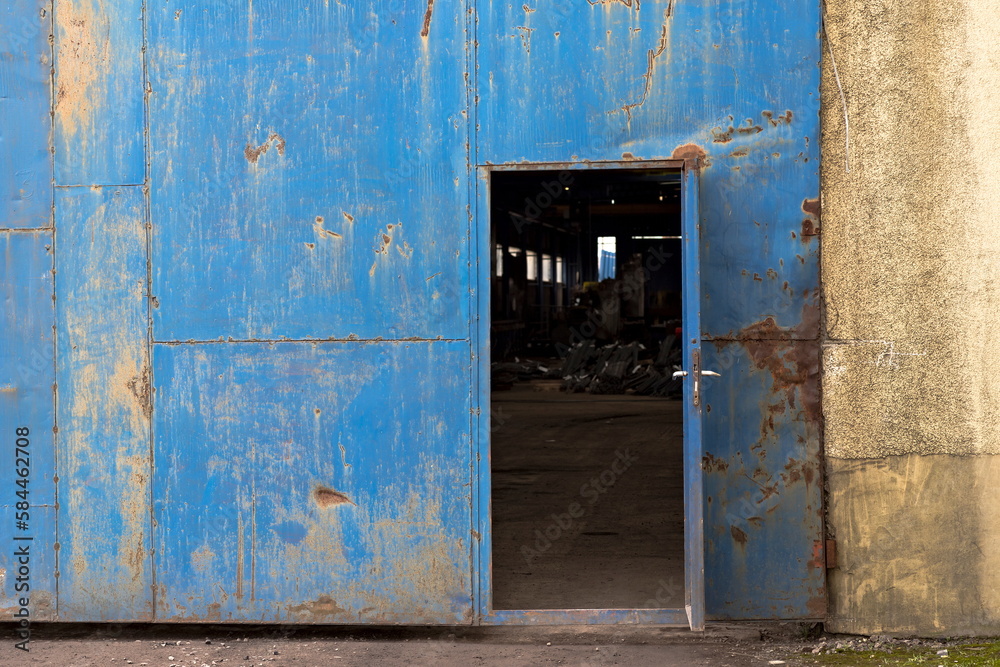 Otwarte drzwi - wmontowane w dużą bramę - do hali przemysłowej . Pordzewiałe pomalowane na niebiesko . W środku widać okna oraz jakieś stalowe elementy konstrukcyjne .
