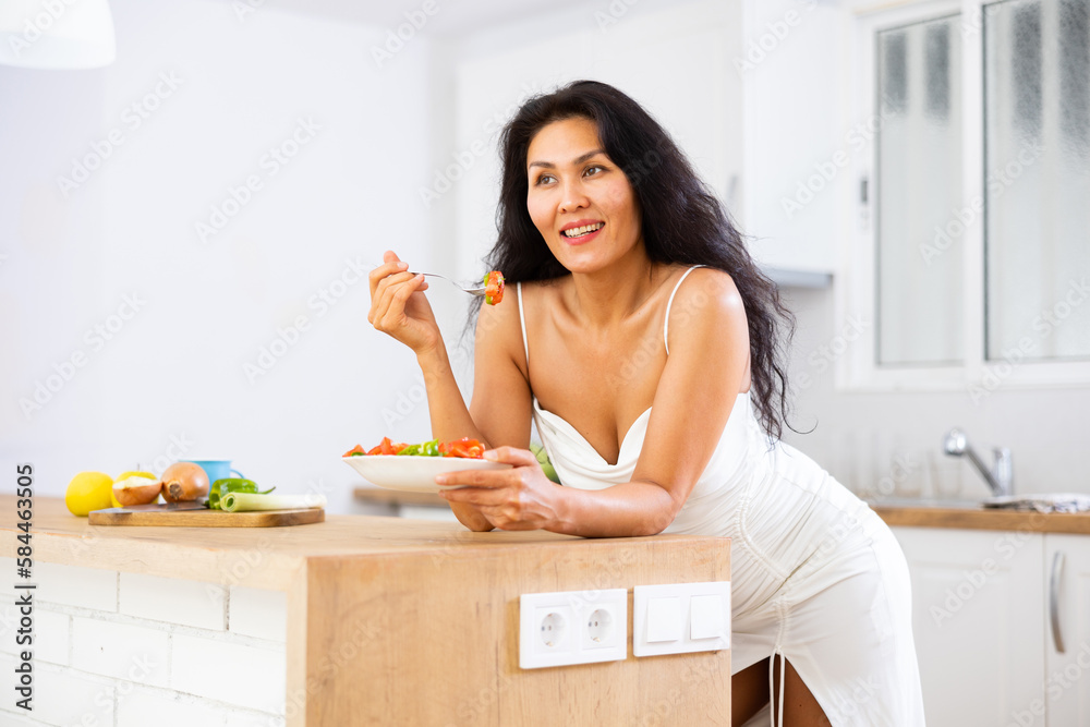 Portrait of cheerful asian woman leaning on table in kitchen and eating salad. Woman having breakfast with fresh salad.