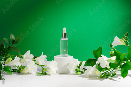 Facial serum in dropper bottle on white podium with freesia flowers  leaves and shadows on green background. Natural skin care cosmetic mockup. Beauty product on pedestal for branding or presentation.