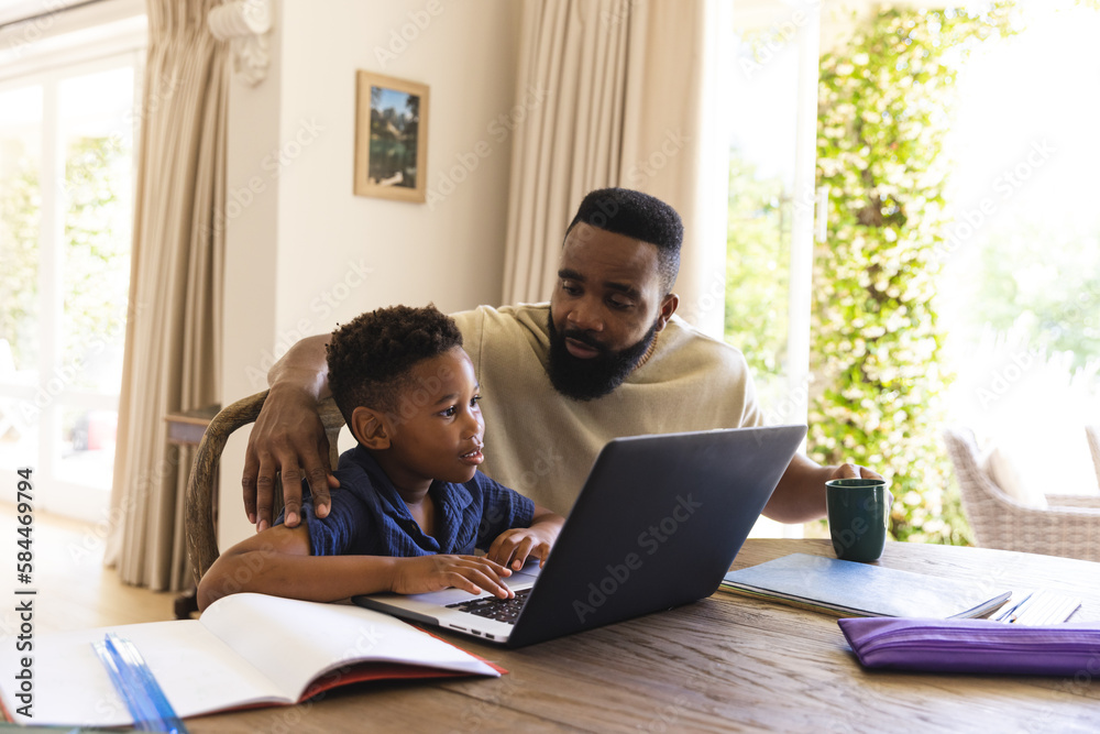 Happy african american father and son sitting at table, doing homework together, using laptop Happy african american father and son sitting at table, doing homework together, using laptop