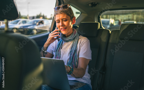 one mature woman caucasian female sitting on the back seat of the car working on laptop computer make a phone call talk in summer day with short gray hair modern on road wearing casual copy space