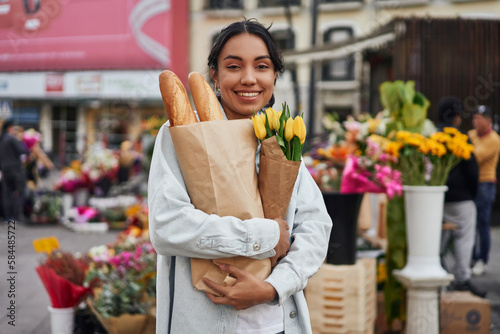 Young Latina woman smiling while buying yellow tulips and bread from a street vendor's stall, adding warmth to her day.