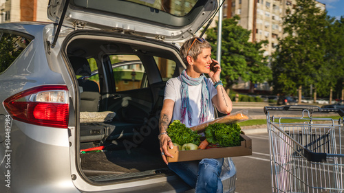 One woman mature caucasian female sit in the back trunk of her car on the parking lot of the supermarket shopping mall or grocery store with vegetables food in box putting them in the vehicle