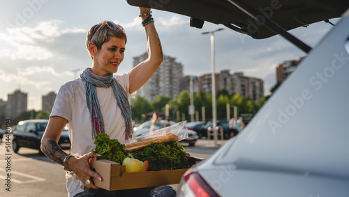 One woman mature caucasian female stand at the back trunk of her car on the parking lot of the supermarket shopping mall or grocery store with vegetables food in box putting them in the vehicle