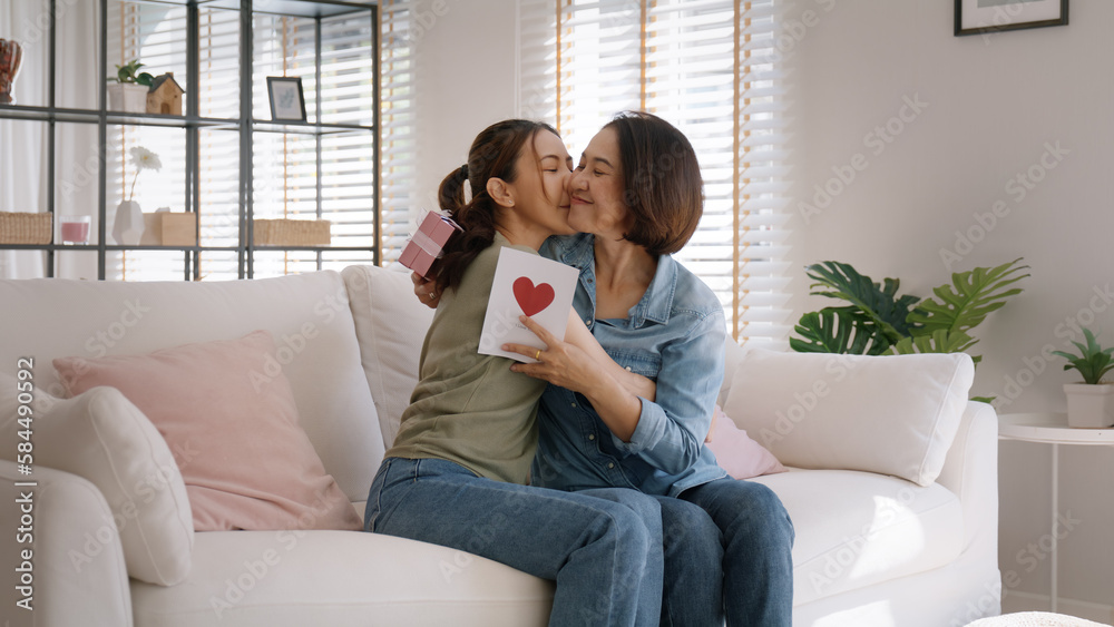 Fototapeta premium May Mother's day young adult grown up child cuddle hug give flower gift box red heart card to mature middle aged mum. Love kiss care mom asia people sitting at home sofa happy smile enjoy family time.