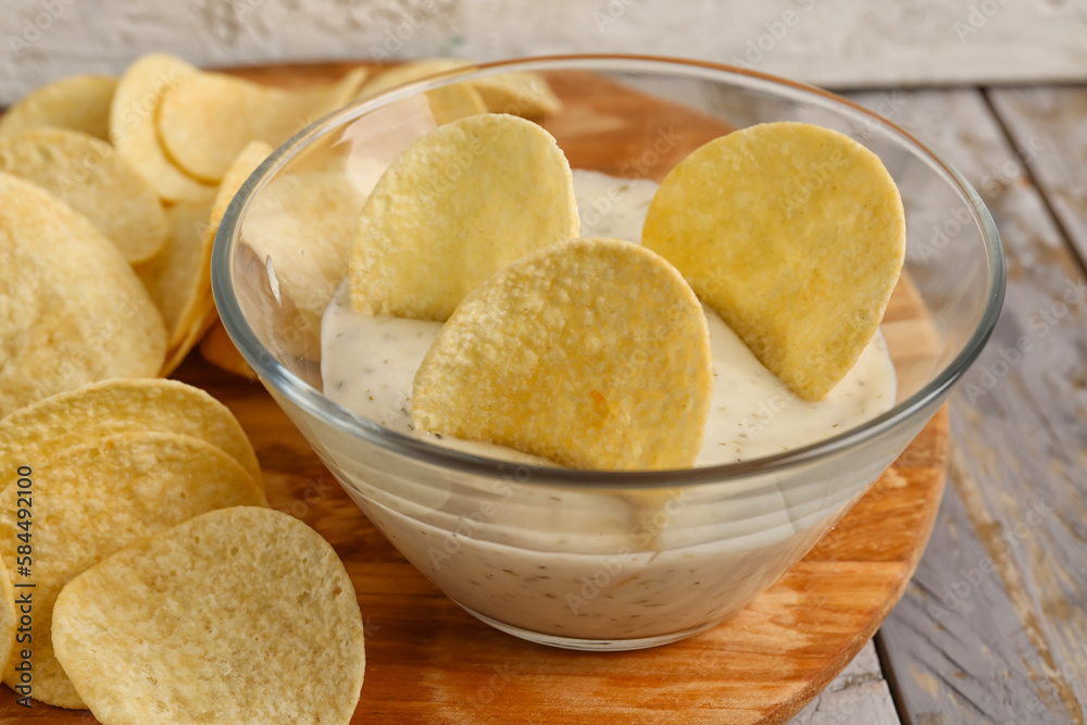 Board with delicious potato chips and bowl of sauce on grey wooden background