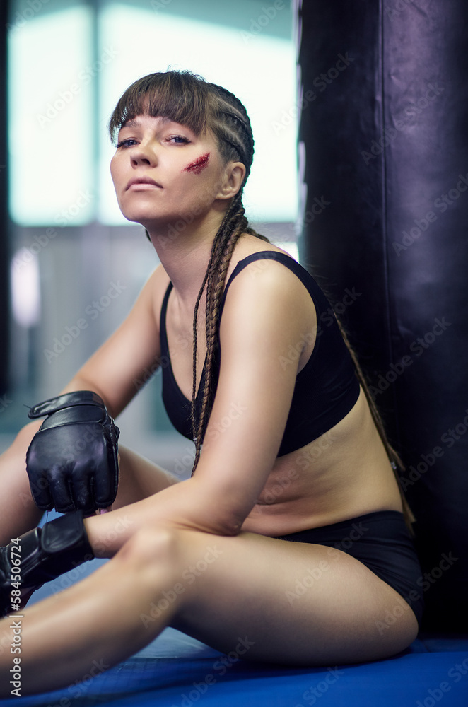 Tired female fighter sitting on the floor leaned against a punching bag in the gym.