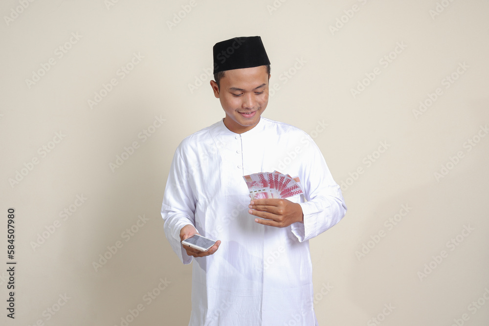 Portrait of attractive Asian muslim man in white shirt showing one hundred thousand rupiah while using mobile phone. Financial and savings concept. Isolated image on gray background Portrait of attractive Asian muslim man in white shirt showing one hundred thousand rupiah while using mobile phone. Financial and savings concept. Isolated image on gray background