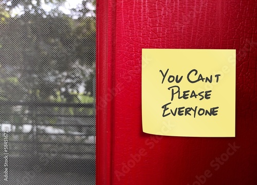 Yellow note stick on red wall with handwritten text You Can’t Please Everyone - self reminder to stop trying to please everyone and be your authentic self