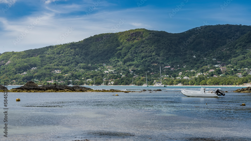 The rocky seabed was exposed at low tide. The boat is moored in shallow water.  Yachts in the distance. A green hilly island against a background of blue sky and clouds. Seychelles. Mahe