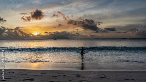 Beautiful tropical sunset. The sun is already low. The clouds are highlighted in orange  pink.  In the ocean there is a silhouette of a man waiting for a rolling wave. Footprints on a sandy beach.