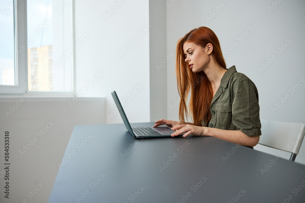 red-haired woman sitting at an empty table works at a laptop in a bright room red-haired woman sitting at an empty table works at a laptop in a bright room