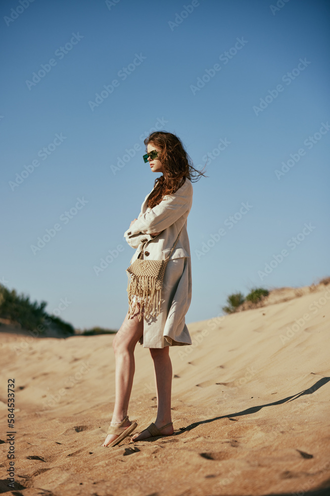 a slender woman in a light long jacket, in black glasses, with a bag slung over her shoulder, stands on the sand against the blue sky