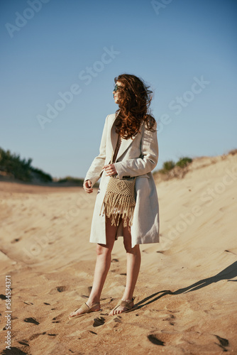 a slender woman in a light long jacket, in black glasses, with a bag slung over her shoulder, stands on the sand against the blue sky