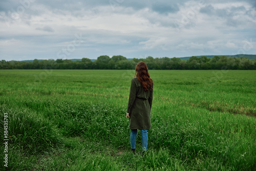 a woman with beautiful, long, red hair stands with her back to the camera in a green field in rainy, spring weather in a long raincoat