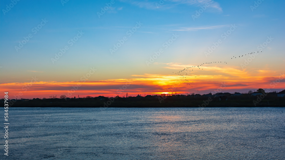 Colorful sunset on a wide river and a flock of migratory birds in the sky