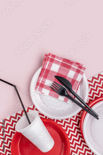 Plastic red and white tableware, paper napkins, pink background, vertical shot. Red and white napkin with a geometric pattern.