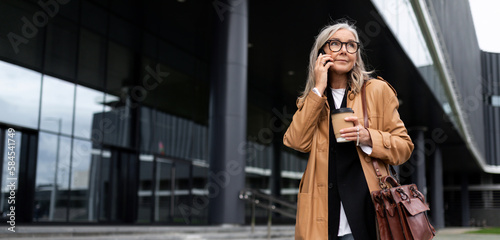 photo of a mature adult female economist with gray hair in a brown coat during a break against the backdrop of a business center