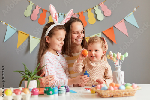 Indoor shot of mother and daughters painting eggs together preparing for Easter against gray decorated wall, infant baby holding colored egg, family spending time together.