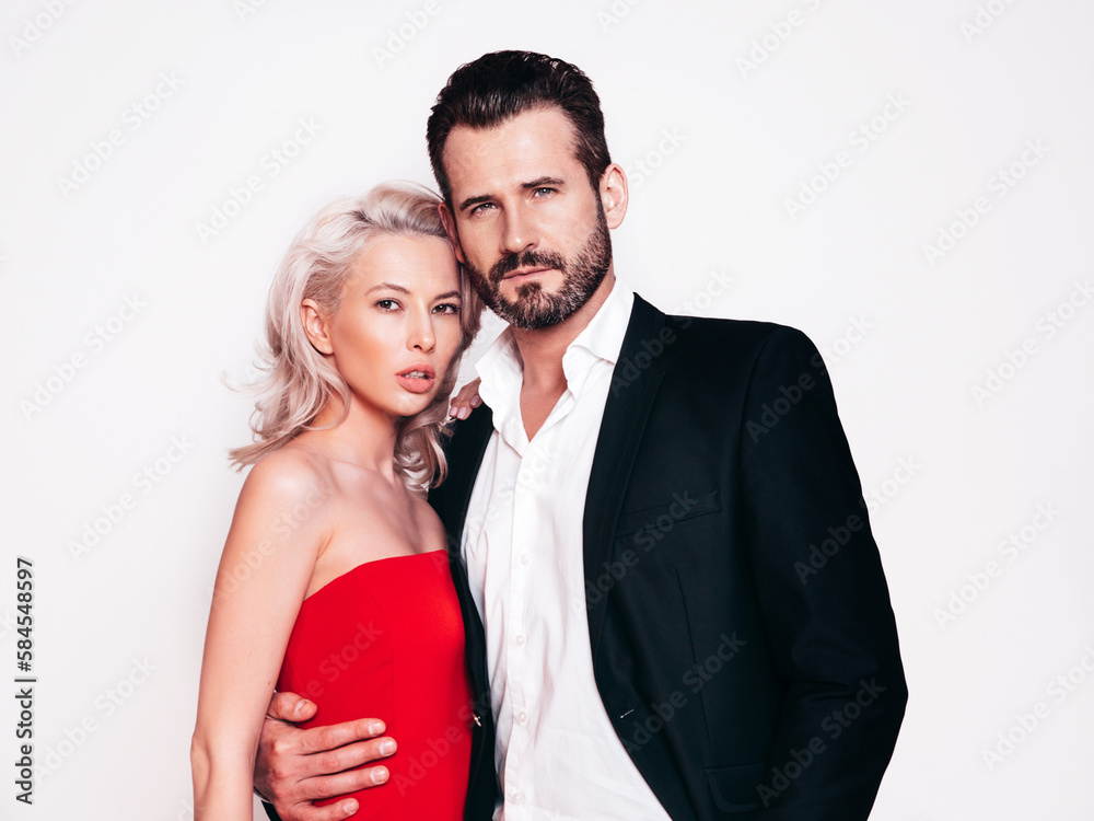 Beautiful fashion woman and her handsome boyfriend in black elegant suit. Sexy blond model in red evening dress. Fashionable couple posing in studio. Brutal man and his female. Isolated on white