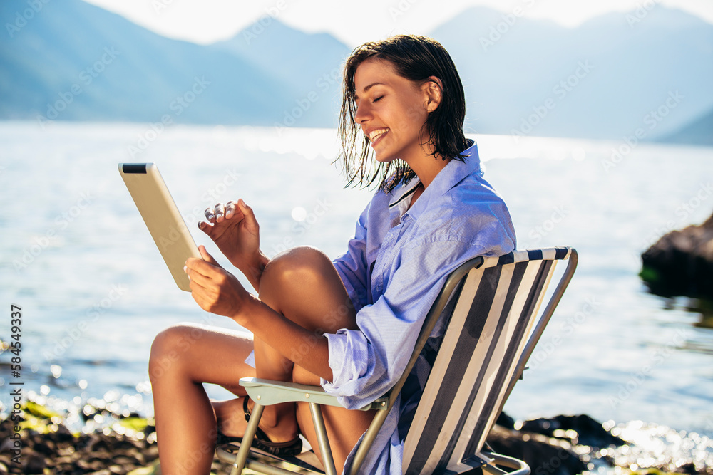 Smiling woman sitting on deck chair by the sea using digital tablet on a sunny day Smiling woman sitting on deck chair by the sea using digital tablet on a sunny day
