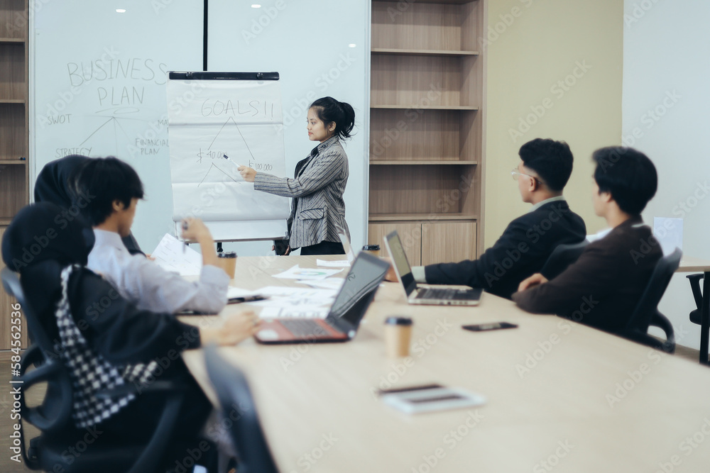 Young beautiful business woman giving flipchart presentation to coworkers during meeting , explain goals strategy at team meeting workshop. Young beautiful business woman giving flipchart presentation to coworkers during meeting , explain goals strategy at team meeting workshop.
