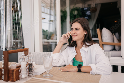 Adorable hispanic brunette young adult woman in white shirt sitting at table at cafe talks by phone with boyfriend looks aside. Pretty caucasian girl excited before date. Summertime. Valentine day.