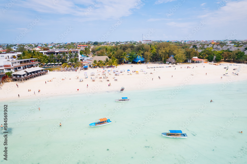 Witness the serene beauty of Zanzibar's tropical coast as fishing boats rest on the sandy beach at sunrise. The top view of the landscape showcases clear blue waters, green palm trees,