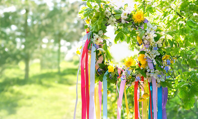 Floral wreath with colorful ribbons on tree in garden, green natural background. floral decor, Symbol of Beltane, Wiccan Celtic Holiday beginning of summer season. witchcraft, folk traditions, rituals