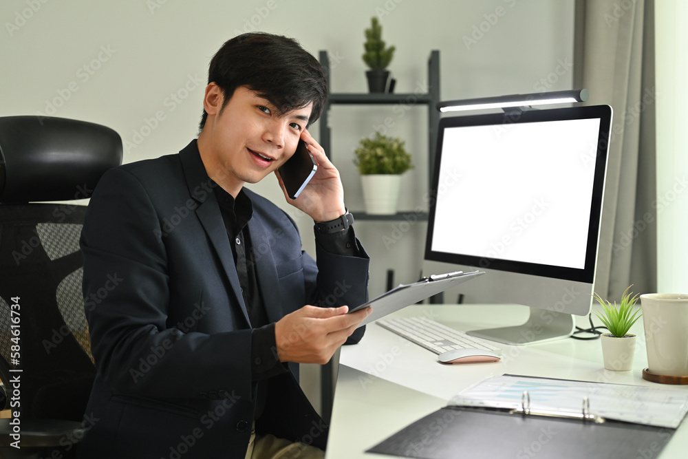 Cheerful asian businessman sitting in front of blank computer screen and talking on mobile phone in modern office Cheerful asian businessman sitting in front of blank computer screen and talking on mobile phone in modern office