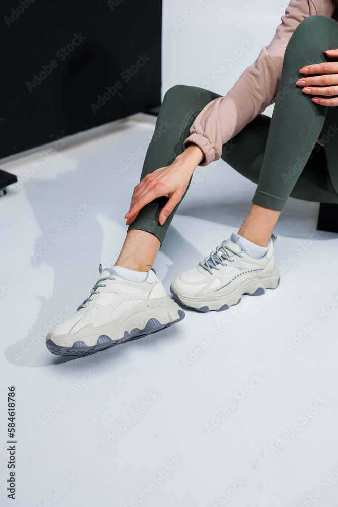 Slender female legs in leggings and white stylish casual sneakers. Women's comfortable summer shoes. Slender female legs in leggings and white stylish casual sneakers. Women's comfortable summer shoes.