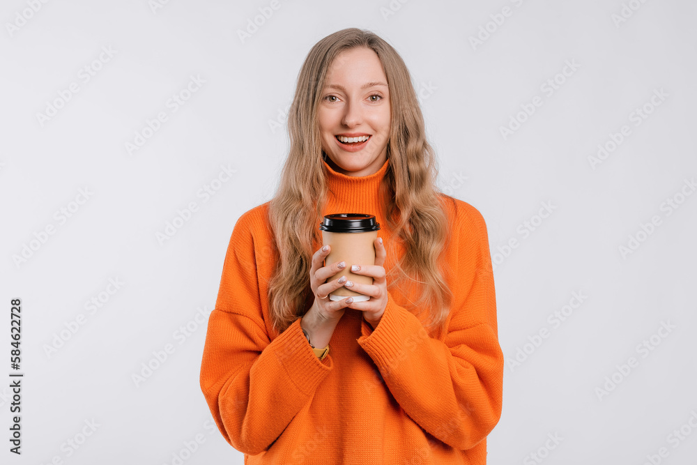 Portrait of a cheerful young woman in an cozy orange sweater holding a paper cup of coffee, standing over neutral background Portrait of a cheerful young woman in an cozy orange sweater holding a paper cup of coffee, standing over neutral background