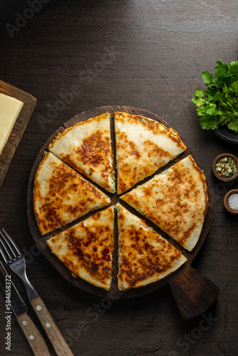 Georgian khachapuri pie with cheese and herbs on a wooden board. Sliced pieces of tortilla stuffed with ingredients on a dark background. Top view, copy space.
