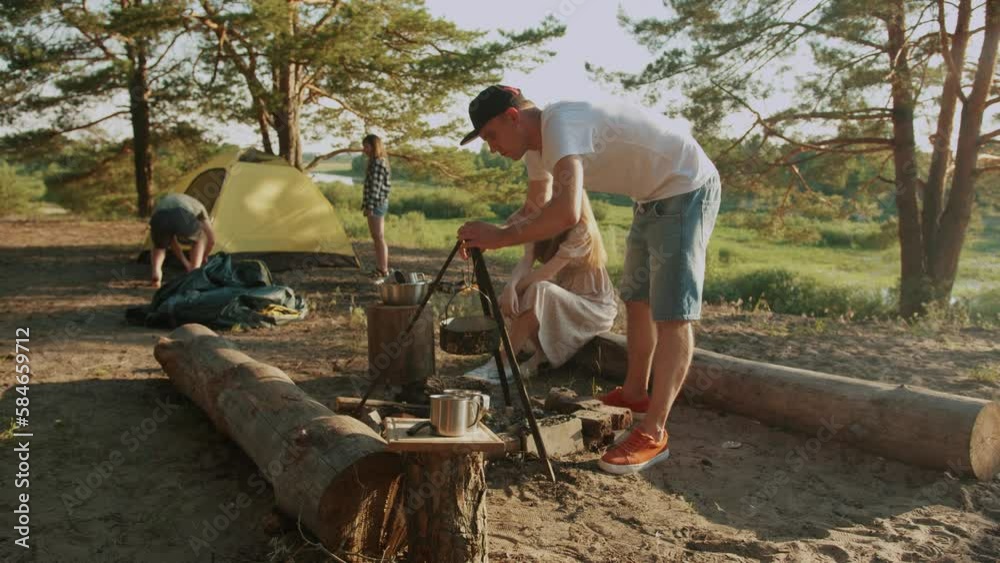 Young guy in cap sets up tripod with a bowler hat over fire while friends set up tent in glade. Slow motion Young guy in cap sets up tripod with a bowler hat over fire while friends set up tent in glade. Slow motion