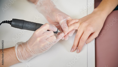 Close up of female manicurist in sterile gloves removing cuticle on woman hand with electric nail drill. Manicure master using cuticle remover device while performing hardware manicure in beauty salon