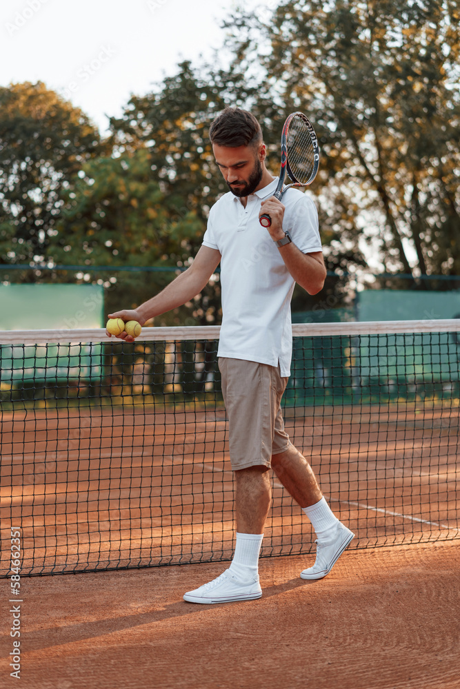 Standing by the net with racket. Young man is on the tennis court at sunny daytime Standing by the net with racket. Young man is on the tennis court at sunny daytime