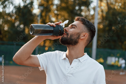 Drinking water, taking a break. Young man is on the tennis court at sunny daytime