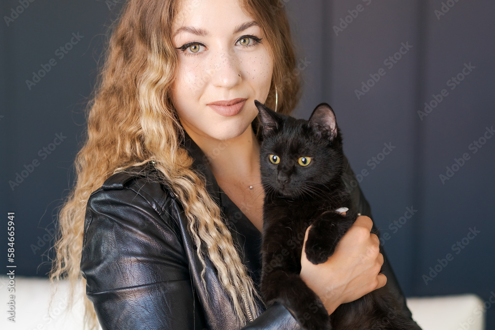 Beautiful young woman in leather jacket holds black cat in her arms near her face, a cute girl with long wavy hair with a pet on a dark background Beautiful young woman in leather jacket holds black cat in her arms near her face, a cute girl with long wavy hair with a pet on a dark background
