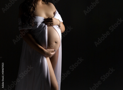 A pregnant woman in a white dress is standing on a black background. Studio pregnancy photo shoot. Copy space. Beautiful pregnancy. Pregnant belly close up.