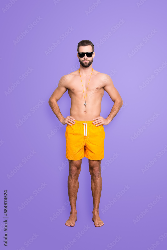 Full size body portrait of muscular athletic lifeguard with stubble in yellow shortsisolated on grey background, holding hands on waist