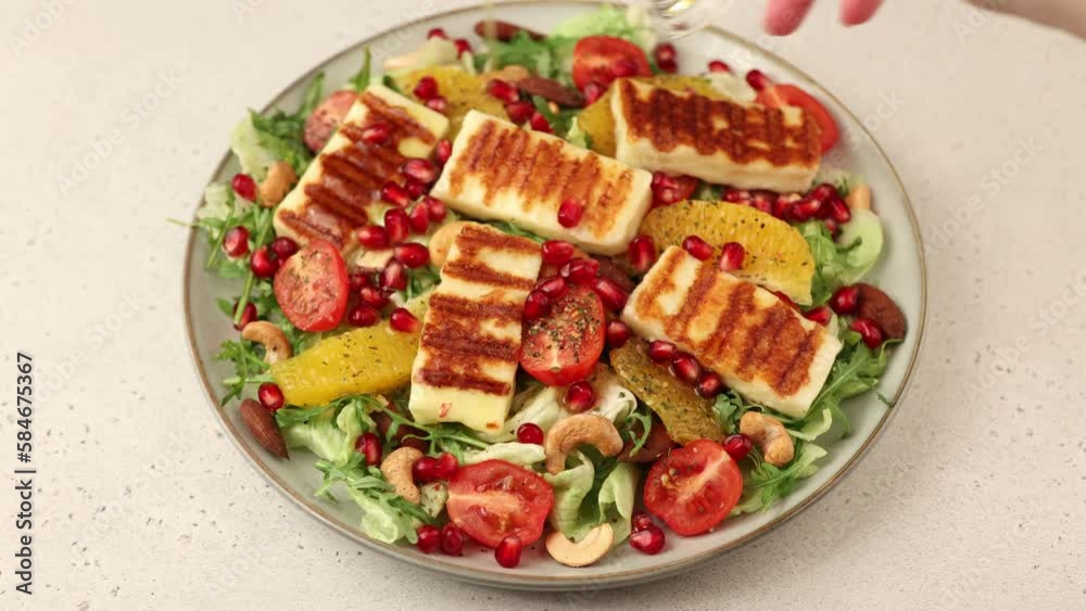 Warm salad with fried halloumi cheese, nuts (cashews and almonds), cherry tomatoes, pomegranate, orange and arugula. Traditional Mediterranean cheese. Woman pouring olive oil on salad. Warm salad with fried halloumi cheese, nuts (cashews and almonds), cherry tomatoes, pomegranate, orange and arugula. Traditional Mediterranean cheese. Woman pouring olive oil on salad.