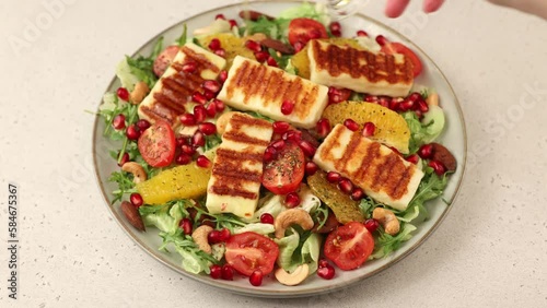 Warm salad with fried halloumi cheese, nuts (cashews and almonds), cherry tomatoes, pomegranate, orange and arugula. Traditional Mediterranean cheese. Woman pouring olive oil on salad. photo