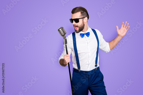 Portrait of handsome attractive singer in blue pants with suspenders and black glasses, singing hit with open mouth in microphone gesture with hand isolated on grey background