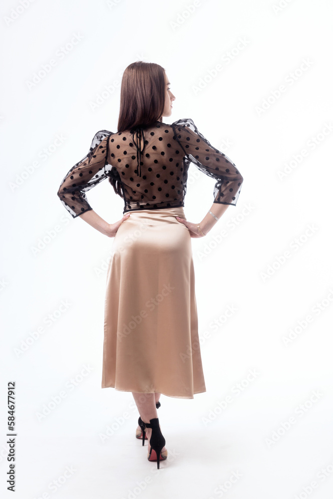 A young woman with dark hair in a black transparent blouse with polka dots and a beige silk skirt posing on a white background with her back.