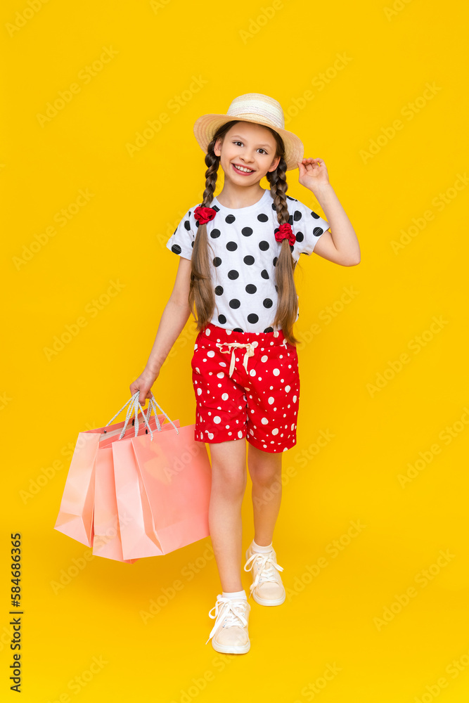 A little girl holds shopping bags. A child smiling in a summer hat and shorts is engaged in children's shopping. A happy child in full growth on a yellow isolated background is holding onto a hat. A little girl holds shopping bags. A child smiling in a summer hat and shorts is engaged in children's shopping. A happy child in full growth on a yellow isolated background is holding onto a hat.