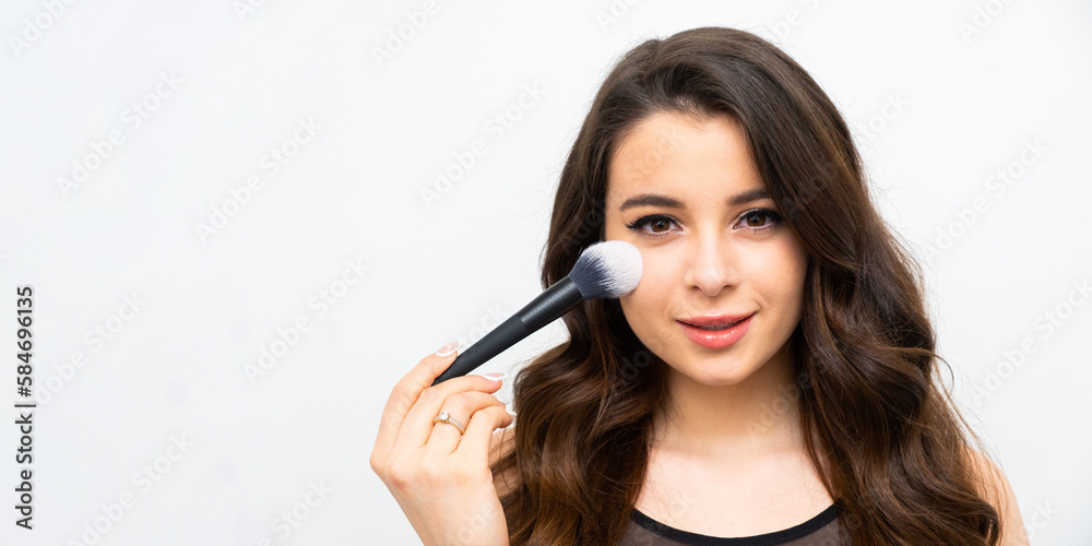 Young woman applies powder on soft facial skin with cosmetic brush. Brunette lady does professional makeup standing on grey background with copy space Young woman applies powder on soft facial skin with cosmetic brush. Brunette lady does professional makeup standing on grey background with copy space