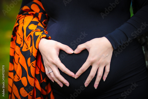 A pregnant woman wearing a black dress holds her stomach with both hands making a sign of love